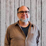 Matthew Callaghan smiles while standing in front of a rustic whitewashed wood plank backdrop, wearing black-framed glasses, a brown button-up overshirt, and a dark crewneck T-shirt. He faces the camera in a professional head-and-shoulders portrait with a closely trimmed gray beard, even lighting, and a friendly, approachable expression.