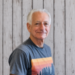 Peter Kostas smiles while standing in front of a rustic whitewashed wood plank backdrop, wearing a gray graphic T-shirt with a colorful landscape design. He faces the camera in a professional head-and-shoulders portrait with short white hair, even lighting, and a friendly, welcoming expression.