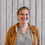 Maya Meislahn smiles while standing in front of a rustic whitewashed wood plank backdrop, wearing clear-framed glasses, a tan corduroy jacket, and a cream blouse with a black geometric pattern. She faces the camera in a professional head-and-shoulders portrait with light brown hair styled up and soft curls framing her face, even lighting, and a warm, welcoming expression.