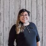 Marysol Alvarez smiles while standing in front of a rustic whitewashed wood plank backdrop, wearing black-framed glasses and a black long-sleeve top with decorative buttons at the neckline. She faces the camera in a professional portrait with long dark hair featuring blonde highlights, even lighting, and a warm, welcoming expression.