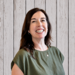 Staff member smiles while standing in front of a rustic whitewashed wood plank backdrop, wearing an olive-green short-sleeve blouse, small hoop earrings, and a delicate pendant necklace. She faces the camera in a professional head-and-shoulders portrait with shoulder-length dark brown hair, even lighting, and a warm, welcoming expression.