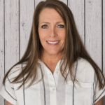 Sandra Goodman smiles while standing in front of a rustic whitewashed wood plank backdrop, wearing a white short-sleeve blouse with gray vertical stripes. She faces the camera in a professional head-and-shoulders portrait with long brown hair, even lighting, and a warm, welcoming expression.
