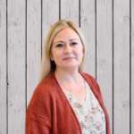 Tammie Wood smiles while standing in front of a rustic whitewashed wood plank backdrop, wearing a rust-colored knit cardigan over a white floral blouse. She faces the camera in a professional head-and-shoulders portrait with long blonde hair, even lighting, and a warm, welcoming expression.