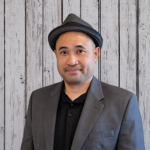 Aristotle Valencia stands in front of a rustic whitewashed wood plank backdrop, wearing a gray blazer over a black collared shirt and a dark gray fedora. He faces the camera in a professional head-and-shoulders portrait with even lighting and a calm, friendly expression.