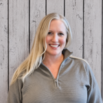 Betsy Nuttall smiles while standing in front of a rustic whitewashed wood plank backdrop, wearing a light gray quarter-zip pullover. She faces the camera in a professional head-and-shoulders portrait with long blonde hair, even lighting, and a warm, welcoming expression.