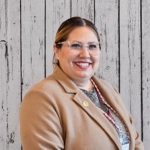Charlene smiles while standing in front of a rustic whitewashed wood plank backdrop, wearing a tan blazer over a patterned blouse, clear-framed glasses, and a beaded lanyard with an ID badge. She faces the camera in a professional head-and-shoulders portrait with dark hair pulled back, even lighting, and a warm, welcoming expression.