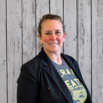 Eo Reveles smiles while standing in front of a rustic whitewashed wood plank backdrop, wearing a black blazer over a gray graphic T-shirt. She faces the camera in a professional head-and-shoulders portrait with her hair pulled back, even lighting, and a warm, welcoming expression.