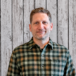 John Shaw smiles while standing in front of a rustic whitewashed wood plank backdrop, wearing a green and gray plaid button-up shirt. He faces the camera in a professional head-and-shoulders portrait with short light brown hair, light stubble, even lighting, and a friendly, approachable expression.