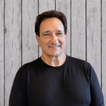 Joseph Guidetti smiles while standing in front of a rustic whitewashed wood plank backdrop, wearing a black crewneck shirt. He faces the camera in a professional head-and-shoulders portrait with dark hair, even lighting, and a warm, welcoming expression.