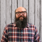 Matt Murdock smiles while standing in front of a rustic whitewashed wood plank backdrop, wearing black-framed glasses and a red, blue, and white plaid button-up shirt. He faces the camera in a professional head-and-shoulders portrait with a bald head, a long full beard, even lighting, and a friendly, welcoming expression.