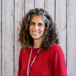 Michelle Kizner smiles while standing in front of a rustic whitewashed wood plank backdrop, wearing a red long-sleeve sweater and a pink lanyard. She faces the camera in a professional head-and-shoulders portrait with shoulder-length curly brown hair featuring gray highlights, even lighting, and a warm, welcoming expression.