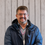 Rich Johnson smiles while standing in front of a rustic whitewashed wood plank backdrop, wearing glasses, a blue Columbia jacket over a gray collared shirt, and a staff lanyard. He faces the camera in a professional head-and-shoulders portrait with short brown hair, a neatly trimmed goatee, even lighting, and a friendly, welcoming expression.